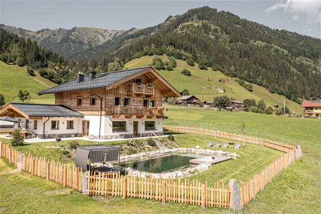 A beautiful wooden house surrounded by green meadows and mountains. In the foreground, there is a pool, surrounded by a wooden fence.