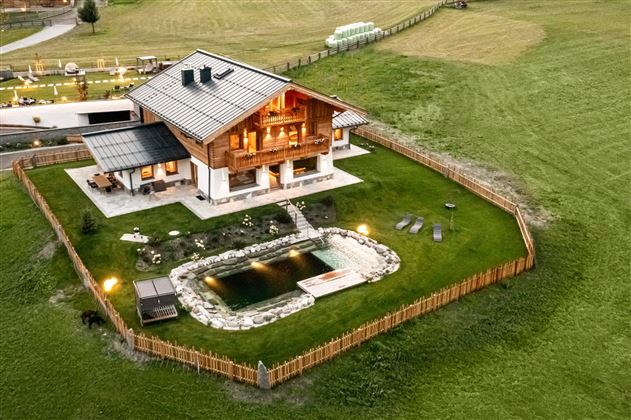 A modern wooden house with a well-maintained garden and a small pond. The surroundings are green and rural, ideal for relaxation.