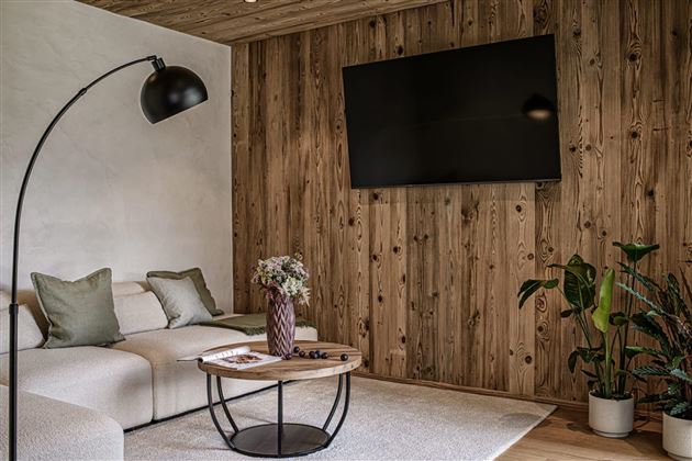 A modern living room with a cozy couch and a coffee table. A large television hangs on the wall, and there are plants for an inviting atmosphere.