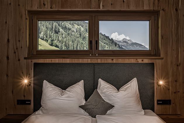 A cozy bedroom with a large bed and pillows. Through the windows, one can see the mountains and the green countryside.