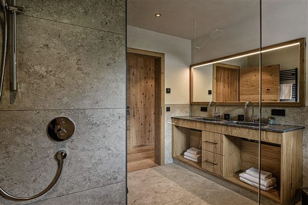 A modern bathroom with an elegant shower and a wooden vanity. The walls are clad in gray tiles and there is a large mirror light.