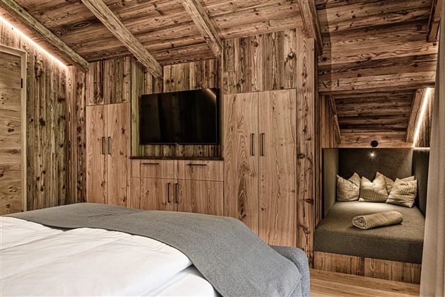 A cozy bedroom with wooden walls and a double bed. A seating area with cushions and a television completes the inviting space.