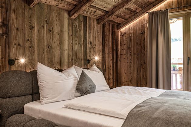 A cozy bedroom with wooden walls and a modern bed.  
Large windows let in daylight and provide access to a balcony.