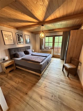 A modern bedroom with a wooden ceiling and floor. There is a double bed, a chair, and a balcony with a view outside.