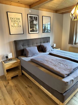 A cozy bedroom with a double bed and wooden floors. Inspirational pictures hang on the wall, and a modern light fixture creates a pleasant atmosphere.