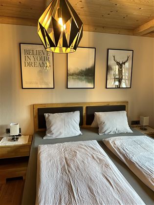 A cozy bedroom with two single beds and bright pillows. Pictures hang on the wall and a modern lamp illuminates the room.