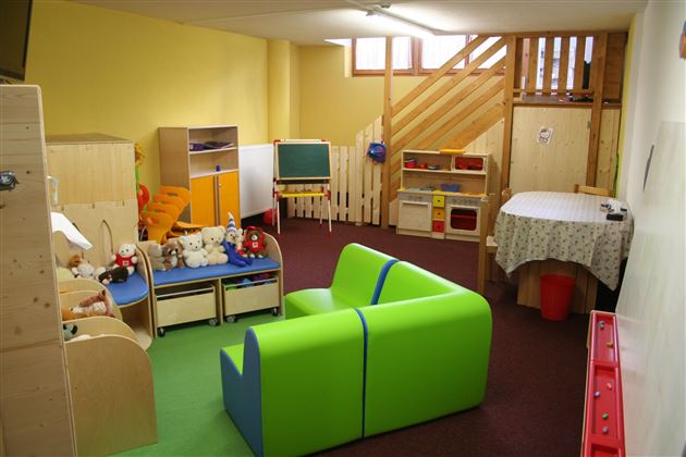 A bright playroom with green and blue furniture. There are toys, a table, and a staircase in the background.