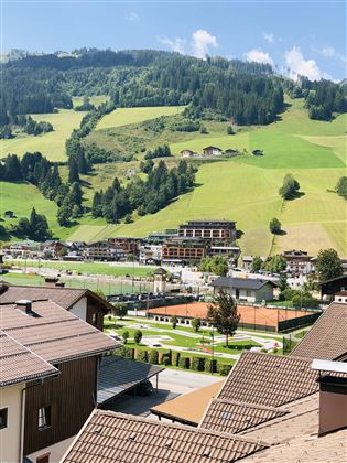 Eine malerische Landschaft mit grünen Wiesen und sanften Hügeln. Im Vordergrund sind einige Häuser und Sporteinrichtungen zu sehen.