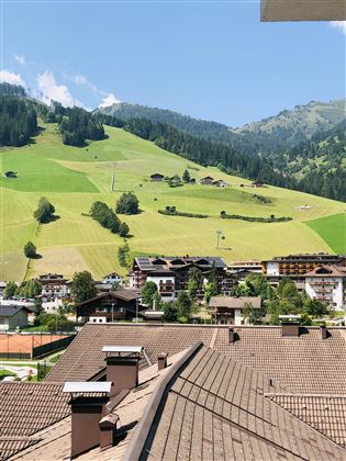 Eine malerische Landschaft mit grünen Wiesen und Bergen im Hintergrund. Im Vordergrund sind zahlreiche Dächer von Häusern und einige Tennisplätze sichtbar.