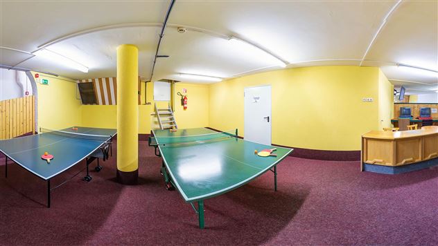 A room with two table tennis tables and yellow walls. In the background, there is a counter and some stairs visible.