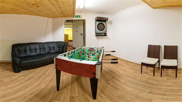 A game room with a foosball table in the center. There is a black sofa and two chairs as well as a dartboard on the wall.