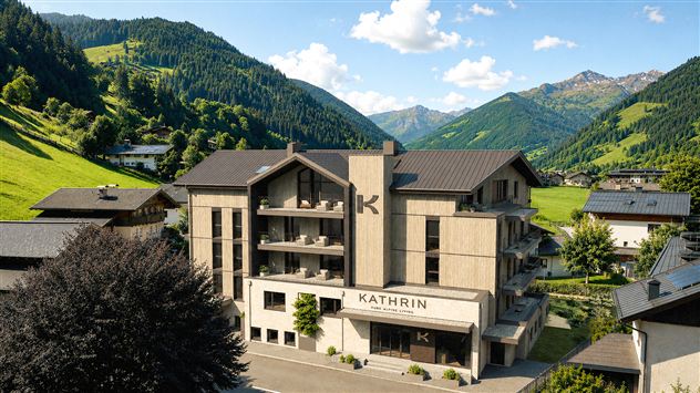 A modern hotel named KATHRIN in the midst of a green mountain landscape. The surroundings show gentle hills and high mountains under a blue sky.