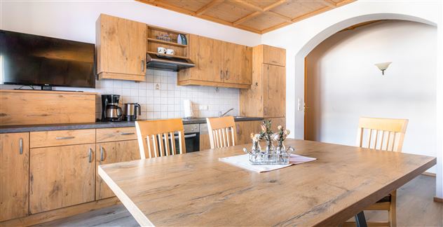 A modern kitchen with wooden cabinets and a large dining table. In the background, there are a stove, a coffee machine, and a television.