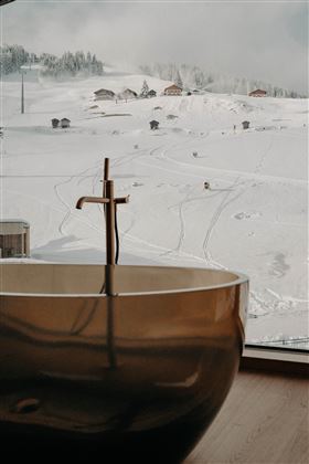 A modern bathroom with an elegant bathtub in the foreground. In the background, a snow-covered landscape stretches out with cottages and snow-covered slopes.
