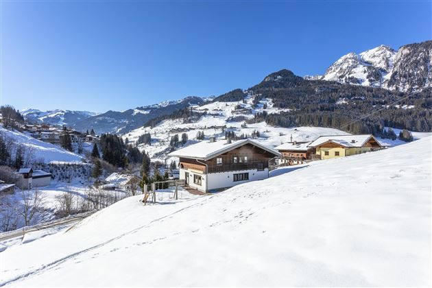 Eine verschneite Winterlandschaft mit Bergen im Hintergrund. Im Vordergrund sind einige traditionelle Holzhäuser zu sehen.