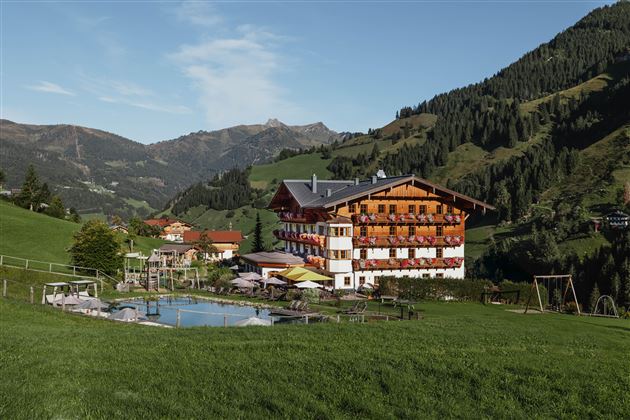 Ein malerisches Hotel inmitten grüner Wiesen und Berge. Im Vordergrund befindet sich ein Schwimmteich und Spielmöglichkeiten für Kinder.