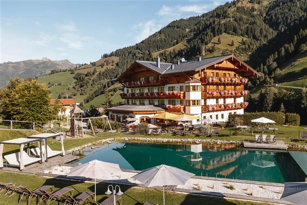 A picturesque hotel in the mountains with a pool and sun loungers in the foreground. Surrounded by green meadows and beautiful landscapes.