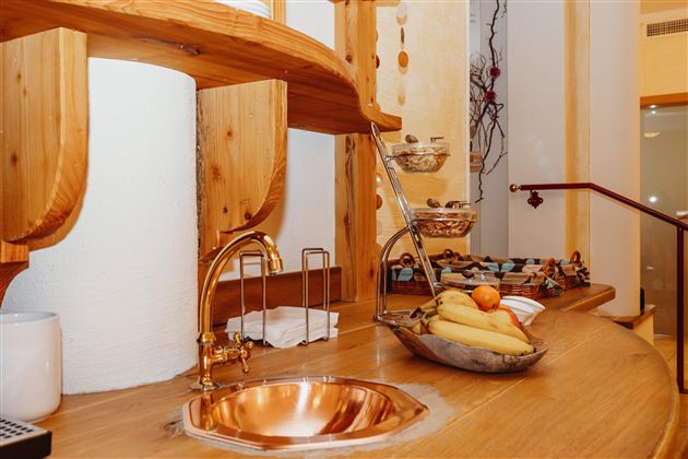 Eine moderne Küche mit einem hölzernen Arbeitsplatz und einer kupfernen Spüle. Im Vordergrund befindet sich eine Schale mit frischem Obst, darunter Bananen und eine orangefarbene Frucht.