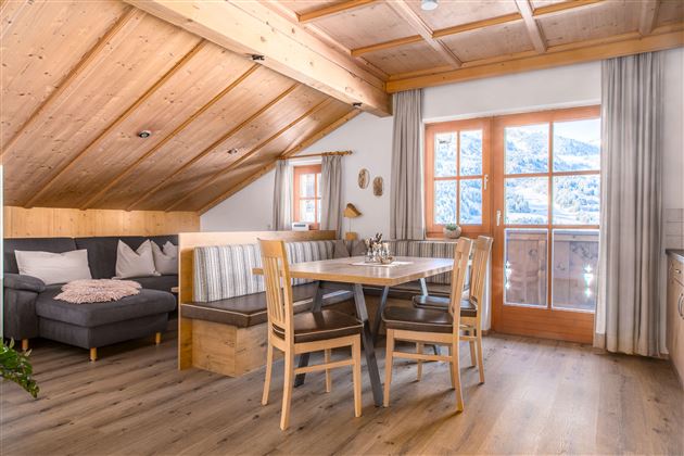 A cozy living kitchen in the attic with a wooden ceiling and a dining table. The large windows offer a beautiful view of the mountains.