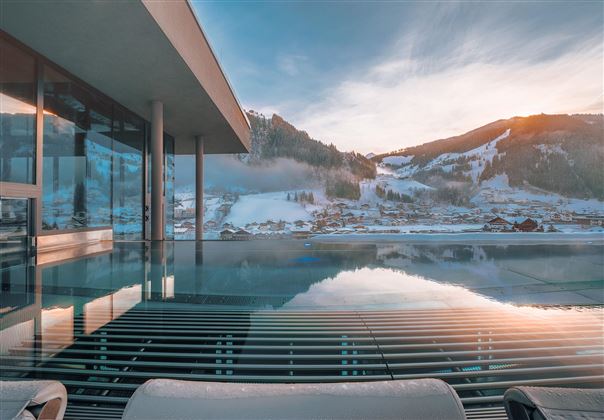 Ein modernes Haus mit großen Fenstern und einem Infinity-Pool. Im Hintergrund sind schneebedeckte Berge und ein klarer Himmel zu sehen.