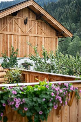 Ein traditionelles Holzhaus mit einem gepflegten Balkon. Im Vordergrund blühen lila Blumen, während im Hintergrund Bäume und Berge sichtbar sind.