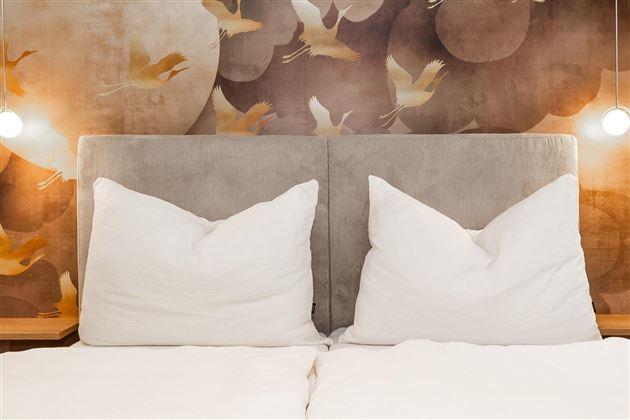 A cozy bed with large, white pillows in front of a stylish wallpaper. The soft lighting creates a relaxed atmosphere.