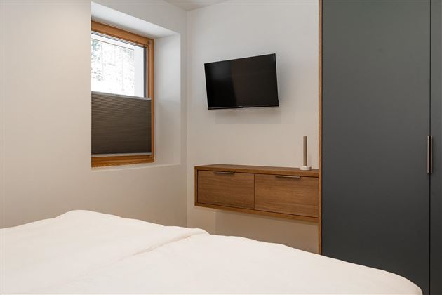 A modern bedroom with a large window and a TV on the wall. The furniture is simple and functional, with a wooden wardrobe and a double bed.