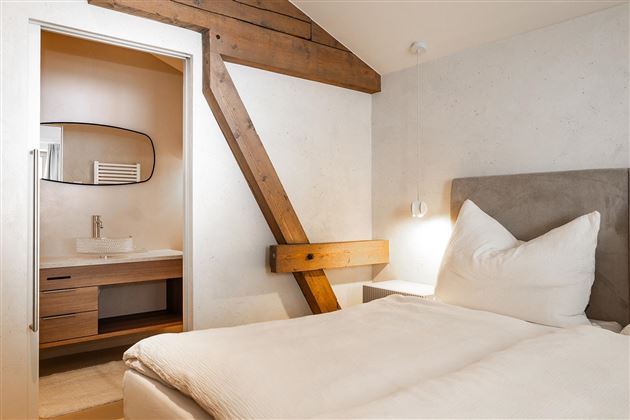 A cozy bedroom with a large bed and modern furniture. In the background, a bathroom with a sink is visible.