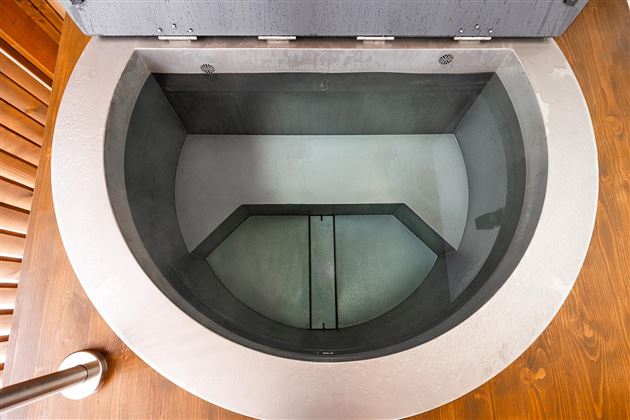 A round, modern water tank made of steel with a clear, transparent bottom. The tank is placed on a wooden floor and has a minimalist and clean appearance.