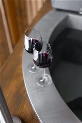 Two wine glasses with red wine are positioned at the edges of a whirlpool. In the background, a wooden structure is visible.