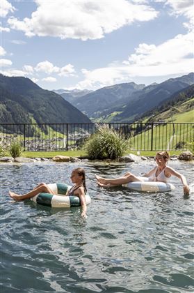 Two people are relaxing in a pool with a view of the mountains. The sky is clear, and the landscape is green and hilly.