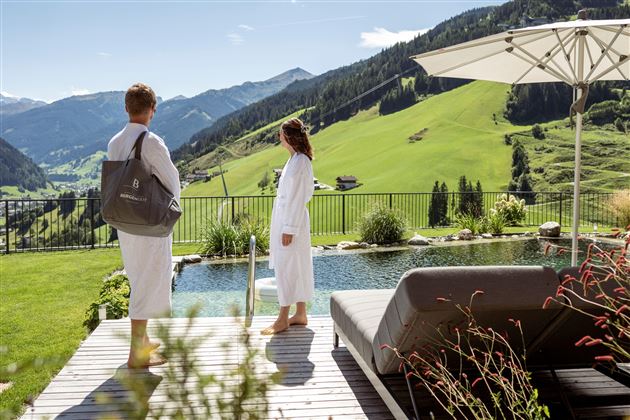 A relaxing spa atmosphere with a view of green mountains and a tranquil landscape. Two people in bathrobes enjoy the view next to a pool.