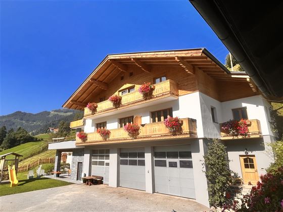 Ein schönes Ferienhaus im alpinen Stil mit blühenden Balkonkästen. Im Hintergrund sind grüne Hügel und ein blauer Himmel zu sehen.