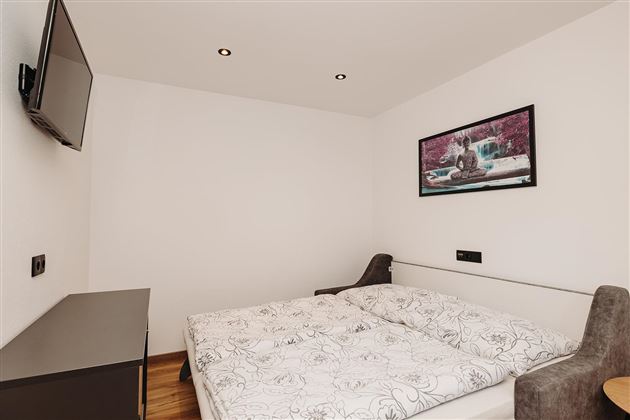 A modern bedroom with a double bed, a television, and a table. The walls are white, and a picture hangs above the bed.