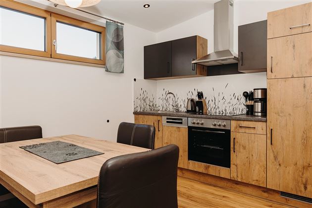 A modern kitchen with wooden cabinets and a dining table. The amenities include a stove, a coffee maker, and large windows for plenty of light.