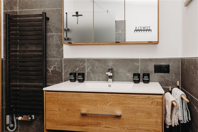 A modern bathroom with a wooden sink vanity and mirror cabinets. The walls are in a gray shade, and towels as well as a towel holder are visible.