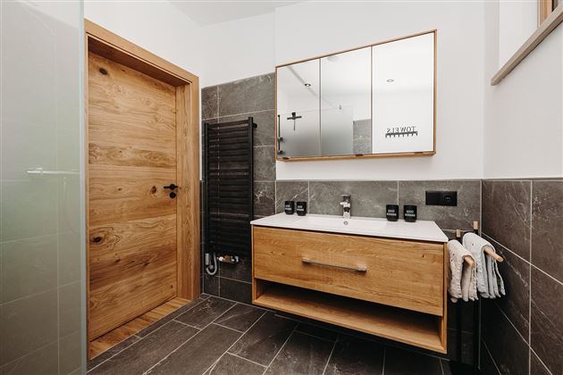 A modern bathroom with a wooden shower door and an elegant wooden sink. The dark tiles and the large mirror wall create a stylish atmosphere.