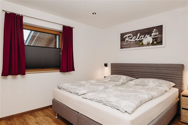 A cozy bedroom with a large bed and soft blankets. Red curtains and a picture with the word "Relax" create a relaxing atmosphere.