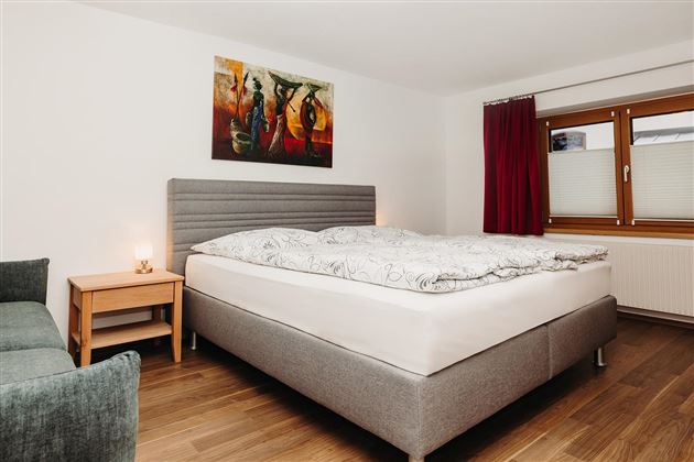 A modern bedroom with a large bed and a cozy seating area. A colorful painting hangs on the wall, and the room has large windows with curtains.