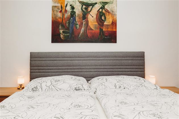 A cozy bedroom with a gray headboard and bed linen with a floral pattern. A colorful piece of artwork hangs on the wall, and two bedside lamps provide soft light.