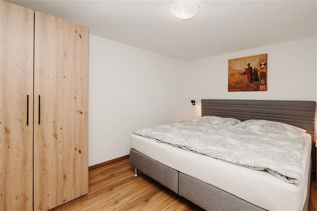 A modern bedroom with a large bed and a wooden wardrobe. The walls are bright and a piece of art hangs above the bed.