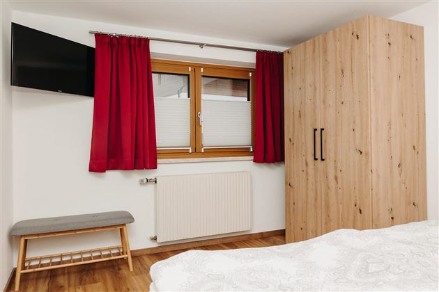 A modern bedroom with a large wooden wardrobe and red curtains. A television is mounted on the wall, and a small stool is next to the heater.