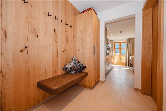 A bright hallway with wooden paneling and a cozy bench. In the background, an open kitchen and a window with a view outside can be seen.