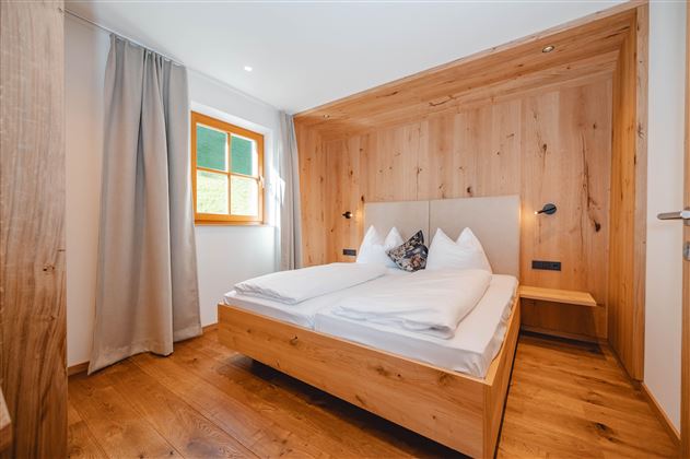 A cozy bedroom with wooden interiors and a large double bed. The window lets in gentle light, creating an inviting atmosphere.