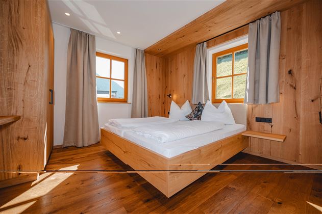 A cozy bedroom with wooden paneling and large windows. The bed is neatly made, and the room feels bright and inviting.