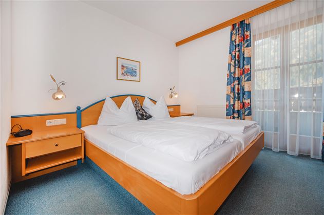 A cozy hotel room with a large double bed and colorful curtains. The room has a table and a desk, as well as natural light from a window.