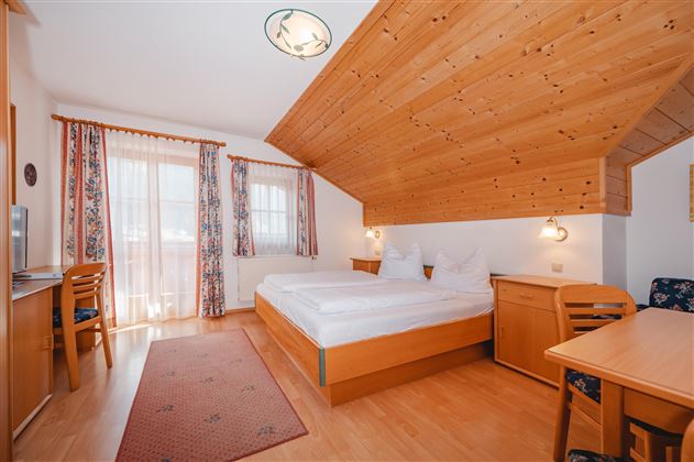 A cozy bedroom with a wooden ceiling and two beds. Large windows let in plenty of daylight and there is a seating area with a table.
