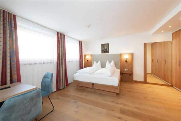 Ein modernes Hotelzimmer mit einem Doppelbett und hellen Vorhängen. Der Boden ist aus Holz und es gibt einen kleinen Schreibtisch sowie einen Kleiderschrank.