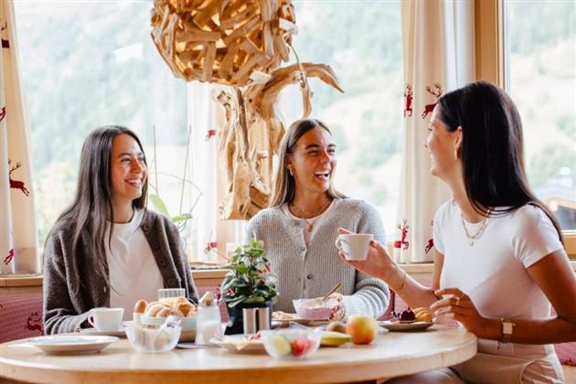 Three women are sitting at the table and enjoying breakfast together. In the background, there are windows overlooking a green landscape.