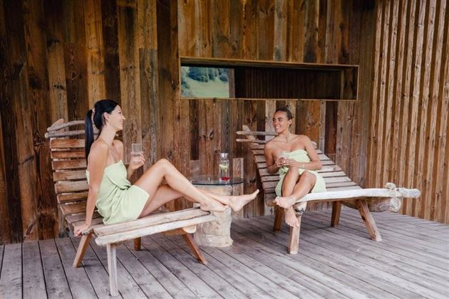 Two women are sitting relaxed on wooden loungers and having a conversation. The background is made of wood and conveys a cozy, natural atmosphere.